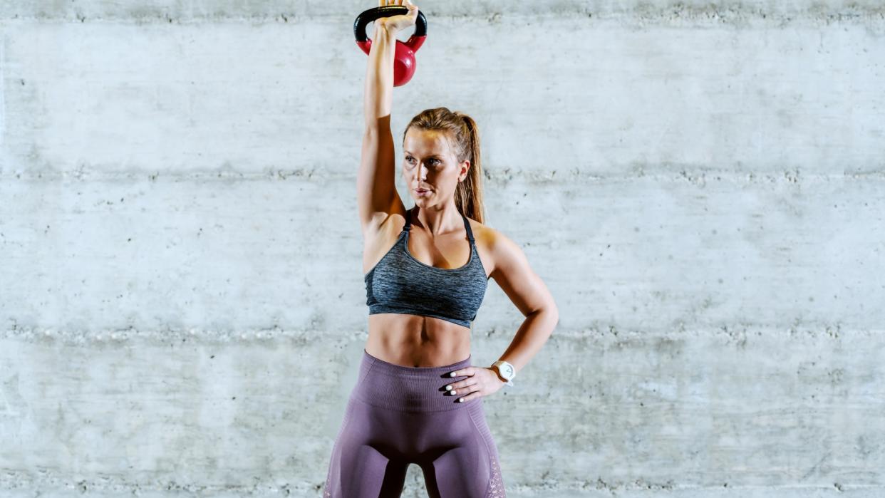  Woman standing against grey wall holding a kettlebell overhead with right arm and left hand on hip. 