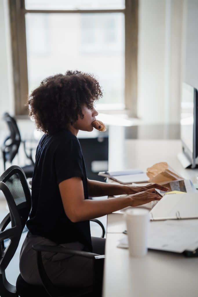 Busy Black woman working at her desk with a donut in her mouth