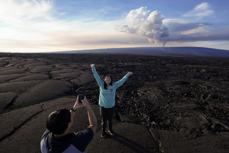 Kelly Ann Kobayashi raises her hands as she poses for a picture for Chad Saito, left, while standing on hardened lava rock from a previous eruption as the Mauna Loa volcano erupts, behind, Wednesday, Nov. 30, 2022, near Hilo, Hawaii. The two were visiting from Honolulu. (AP Photo/Gregory Bull)