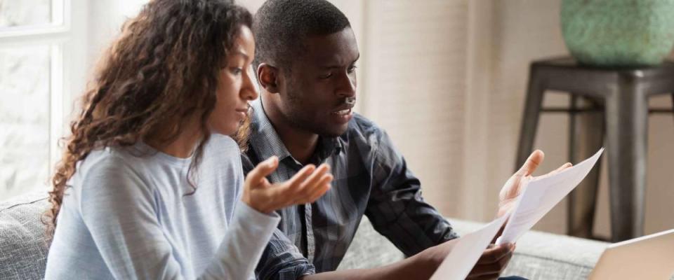 Concerned mixed race husband and wife take care of domestic bills reading papers at home, worried black couple consider bank documents, think of mortgage or loan, check paperwork together