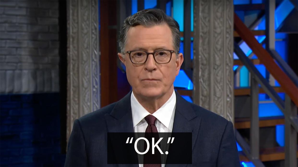 A man in a suit stands on a talk show stage staring at the camera with a neutral expression. The caption below reads: 