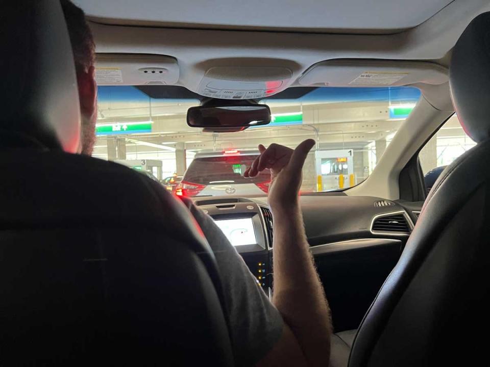 A man driving a car as seen from the backseat as he gives a Hawaiian shaka sign.