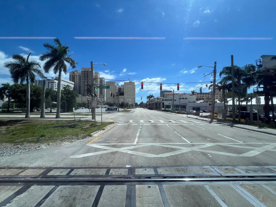 A view looking out of a Brightline train window in Florida. Buildings, palm trees and an empty road are shown.