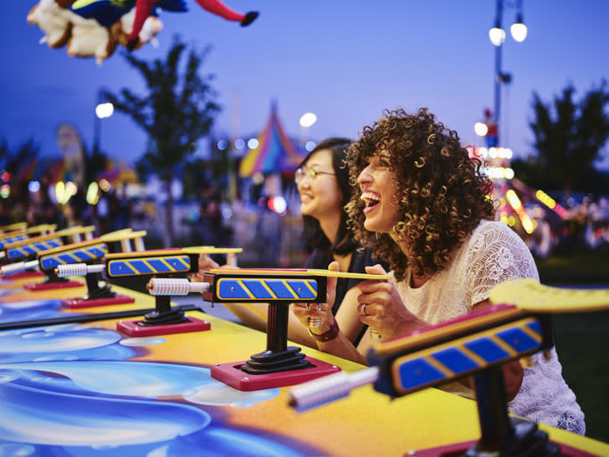 Two women smiling and playing a carnival water gun game at a festive outdoor event. Amusement rides and colorful lights are visible in the background