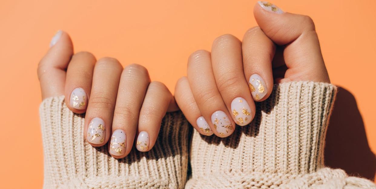 glowing hands in sweater with knitted sleeves and with nude color manicure with gold particles on orange table trendy colors of the year