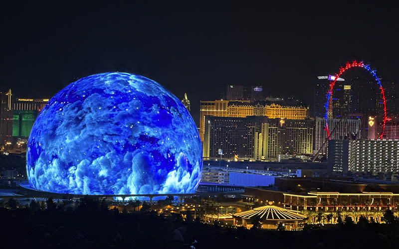 The MSG Sphere illuminates the Las Vegas skyline. The sky is dark showing lit buildings and a Ferris wheel while near the center of the screen is seen a giant blue sphere illuminated with points of light and clouds.