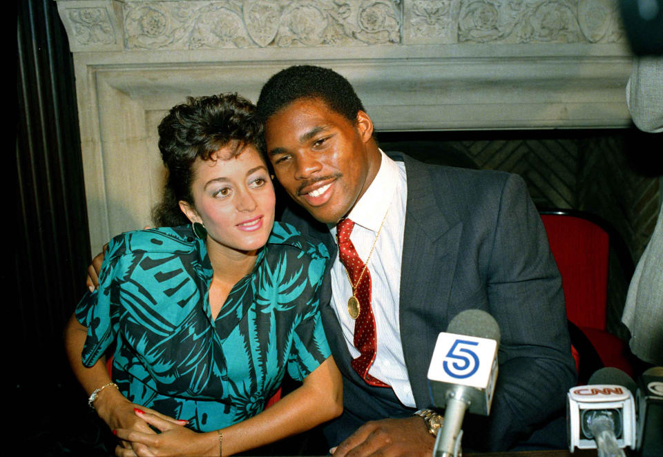 Herschel Walker poses cheek to cheek with his wife, Cindy, in front of a bank of microphones, with an ornate cared mantel behind them..
