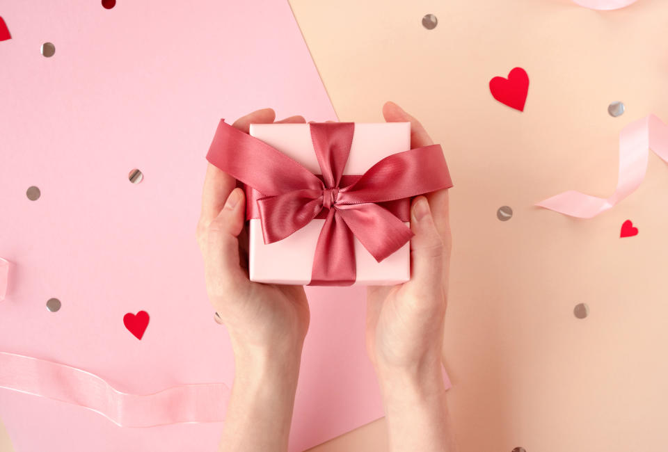 Female hands holding gift box with satin magenta ribbon on festive pink background with confetti and hearts. Valentines Day, International Women Day, Mothers Day Concept