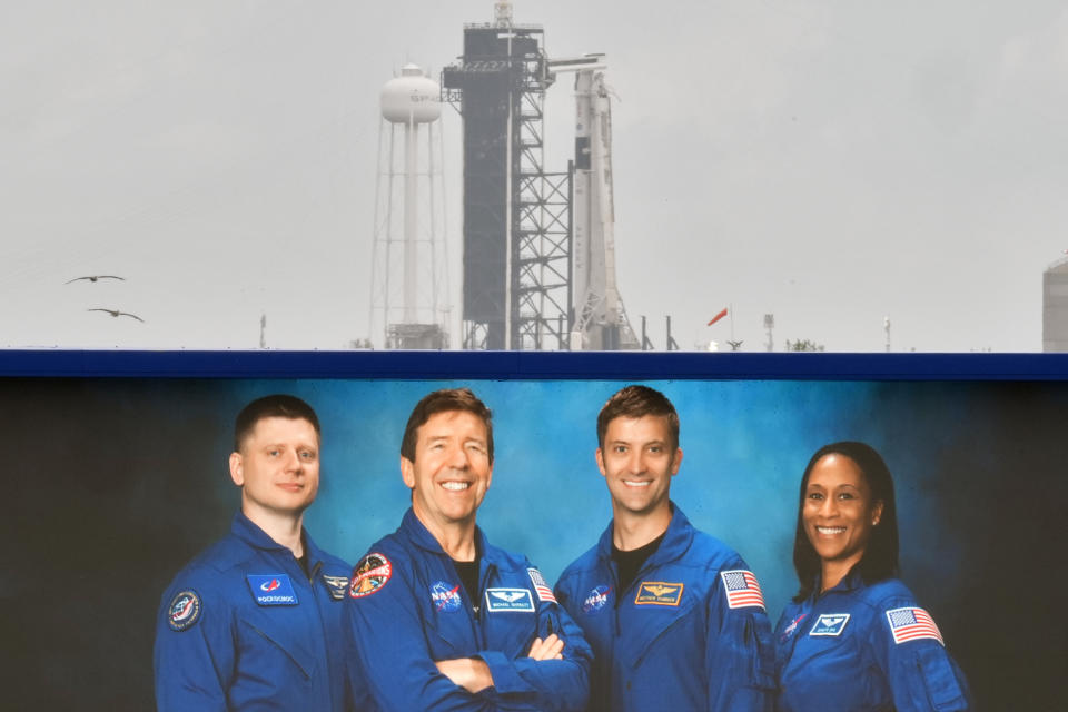 A SpaceX Falcon 9 rocket sits on Launch Pad 39-A, behind a large video photo of the crew, from left, Alexander Grebenkin, of Russia, Michael Barratt, Matthew Dominick, and Jeanette Epps, Friday, March 1, 2024, at the Kennedy Space Center in Cape Canaveral, Fla. Four astronauts are set to lift off Saturday night on a mission to the International Space Station. (AP Photo/Chris O'Meara)