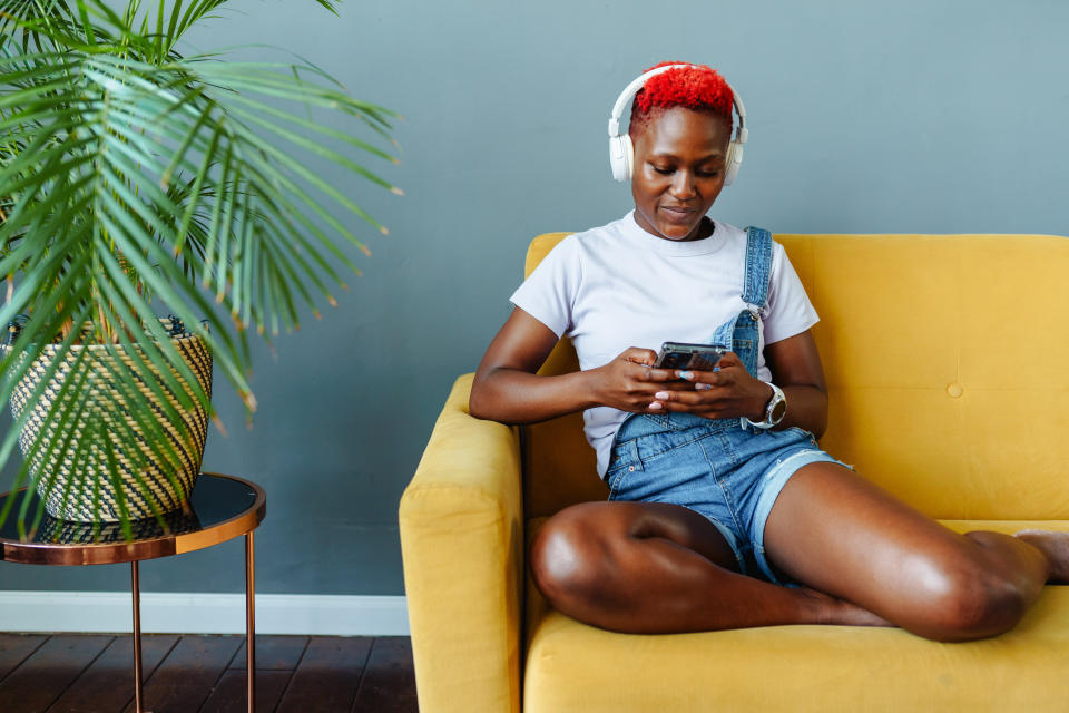 Woman with headphones using smartphone on yellow sofa, relaxed pose, plants nearby