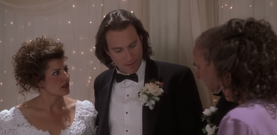 Toula, in a wedding dress, and Ian, in a black tux, stand in front of a wall of fairy lights, looking shocked