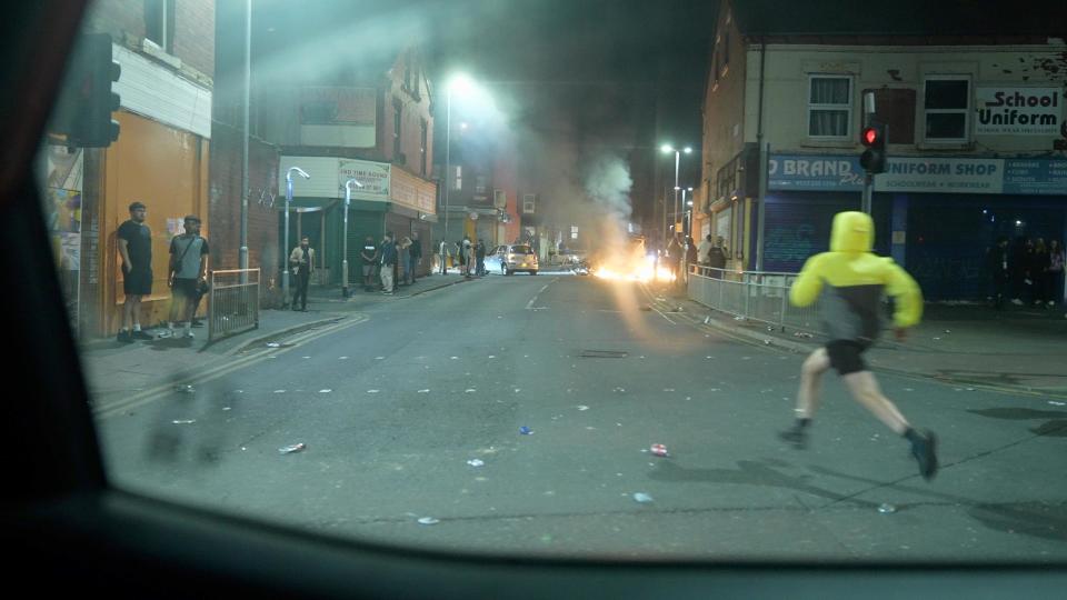 A hooded boy runs across the road with a fire in the background