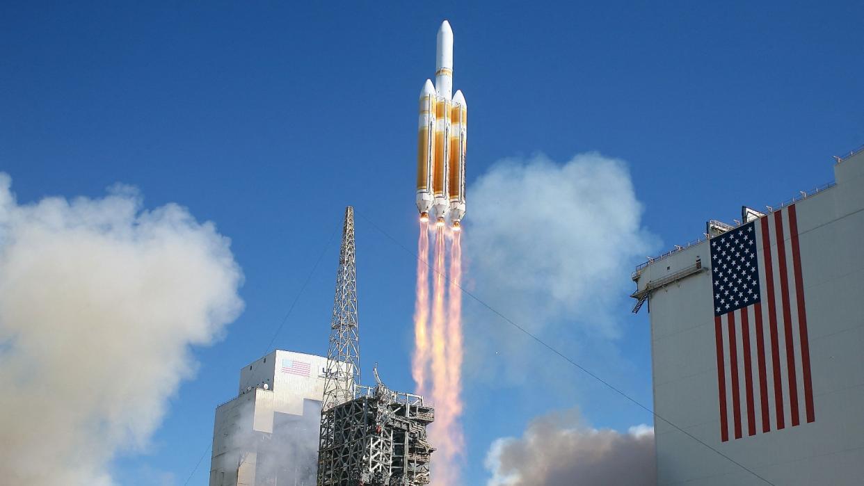  A large white-and-orange rocket lifts off behind an american flag. 