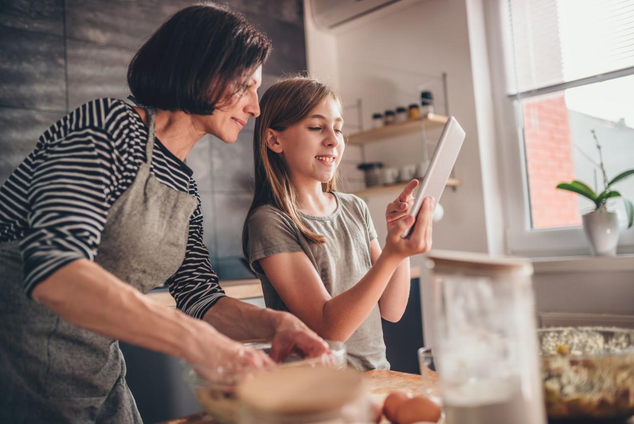 Mother and daughter standing in the kitchen by the wooden table kneading dough and searching apple pie recipe on the tablet