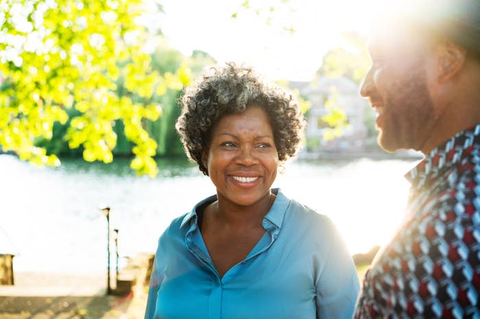 Woman smiling at the camera, man facing her, both by a lakeside with trees in background