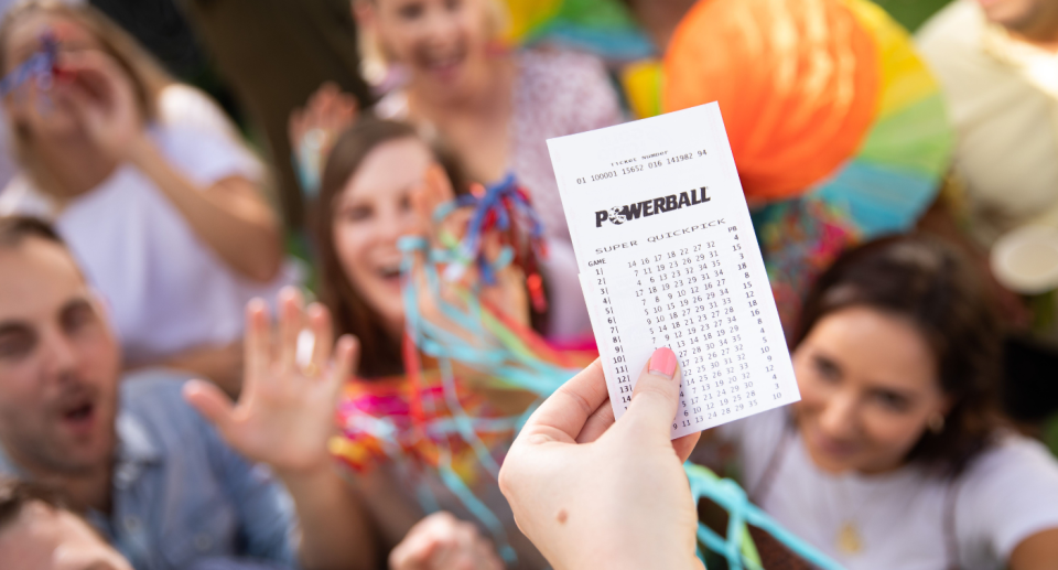 A hand holding a Powerball lotto ticket in front of people celebrating.