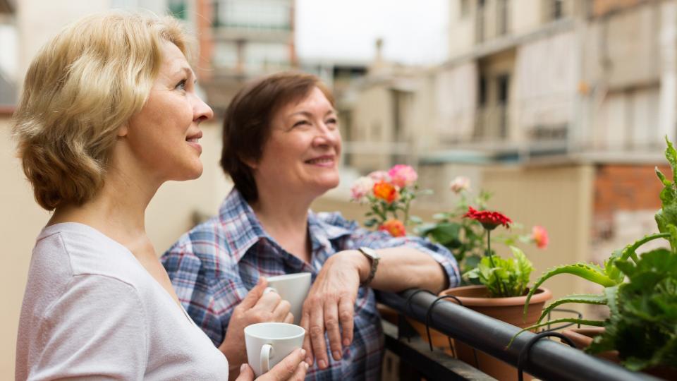 Smiling aged female friends relaxing with cup of coffee on balcony.