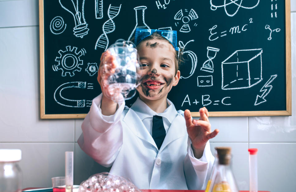 Kid scientist making a mess in a lab with a blackboard in the background filled with science and math drawings.