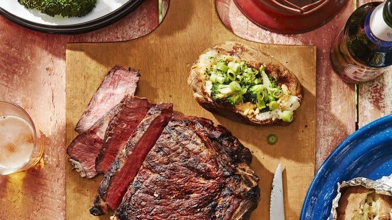 cowboy steak sliced on a wooden cutting board and arranged with potatoes with broccoli and cheddar scallion spread on top