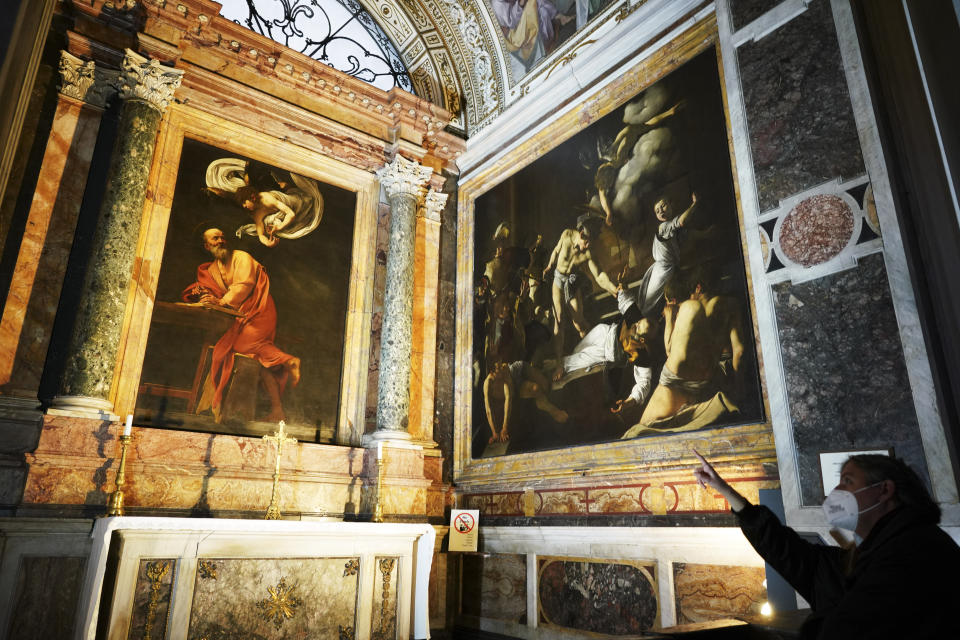 A woman wearing a face mask to curb the spread of COVID-19 points to a cycle of paintings by 16th century artist Michelangelo Merisi, known as Caravaggio, on the life of St.Matthew, inside the Contarelli chapel of San Luigi dei Francesi Church, in Rome, Friday, Dec. 11, 2020. Like elsewhere in Europe, museums and art galleries in Italy were closed this fall to contain the spread of COVID-19, meaning art lovers must rely on virtual tours to catch a glimpse of the treasures held by famous institutions such as the Uffizi in Florence and the Vatican Museums in Rome. However, some exquisite gems of Italy's cultural heritage remain on display in real life inside the country's churches, some of which have collections of renaissance art and iconography that would be the envy of any museum. (AP Photo/Andrew Medichini)