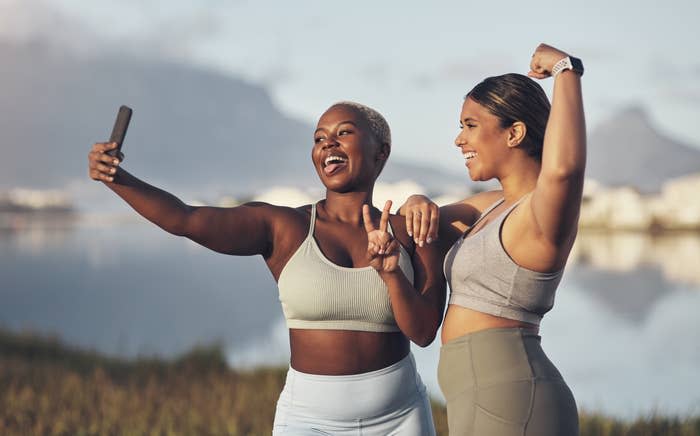 Two women in workout gear take a selfie outdoors by a lake. One flexes her arm muscle while both smile and pose for the photo