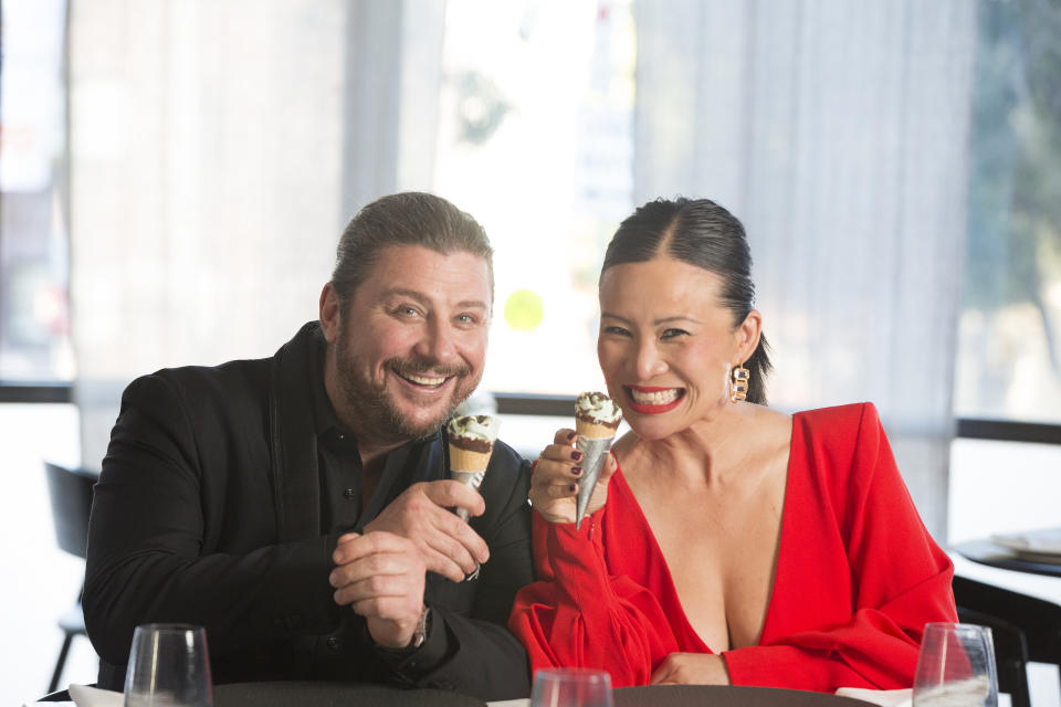 Snackmasters hosts Poh Ling Yeow and Scott Pickett sit at a restaurant dining table while holding mini drumstick ice creams