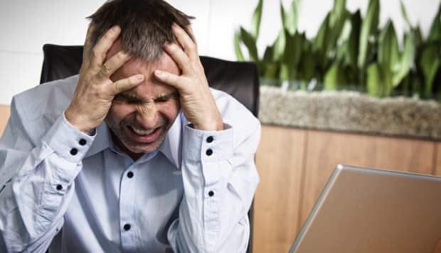 Frustrated overloaded senior businessman being furious about work and clinching his teeth, sitting at office desk in front of la