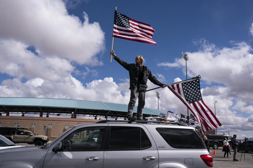 Jeff Cassil waves two American flags from the roof of his car at the beginning of a trucker caravan to Washington, D.C., called the "The People's Convoy," on Wednesday in Adelanto, Calif. A small convoy of truckers demanding an end to coronavirus mandates began a cross-country drive from California to the Washington, D.C., area. (AP Photo/Nathan Howard)