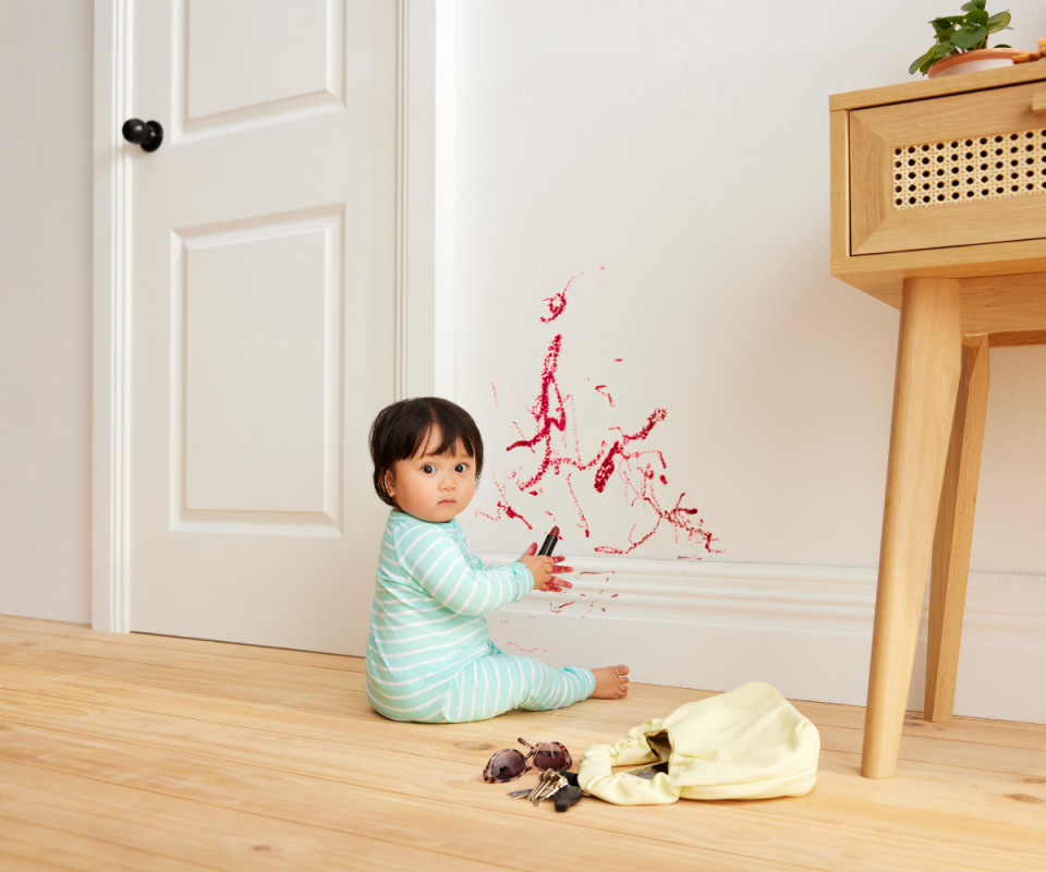 A brown haired baby in a pale green and white stripped onesie sits on a pale timber floor holding red lipstick and draws on a white wall.