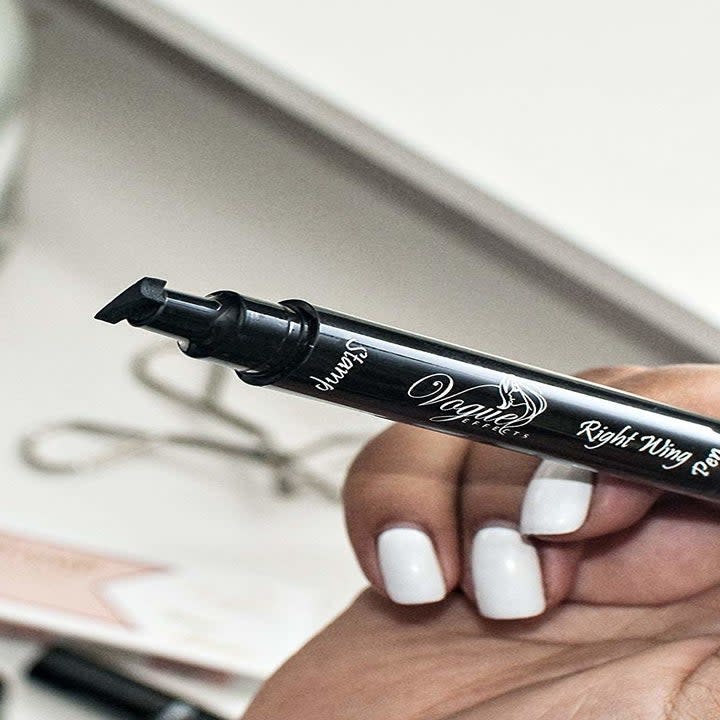 A hand holding the black pen with the winged eyeliner stamp on the end 