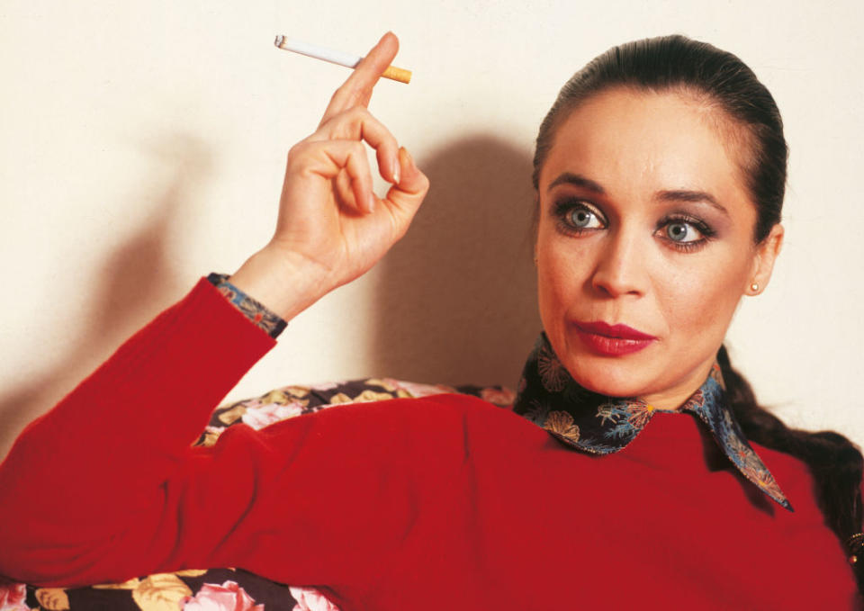 Woman in a floral-collared red sweater holding a cigarette, looking to the side with a serious expression