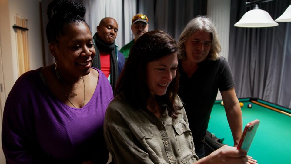 A group of people stand next to a pool table; a smiling woman holds a cellphone as four others look at the screen.