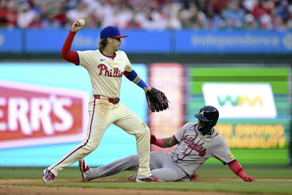 Philadelphia Phillies' Bryson Stott, left, turns a double play on a ball hit by Atlanta Braves' Travis d'Arnaud as Orlando Arcia slides during the fifth inning of a baseball game, Saturday, March 30, 2024, in Philadelphia. (AP Photo/Derik Hamilton)