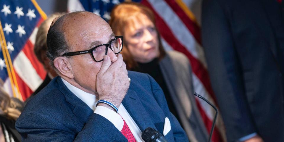 Rudy Giuliani covers his mouth with his hand at a press conference, with American flags visible in the background.
