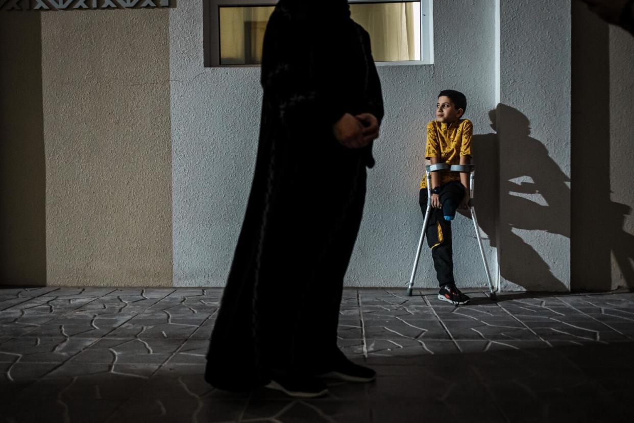 A child with one leg leans on crutches, his shadow falling against a wall, as an adult stands in the foreground.