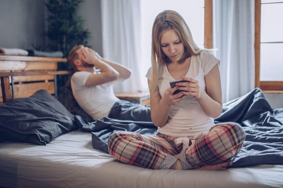 Young man upset while his girlfriend uses a smartphone in bed. Photo: Getty Images.