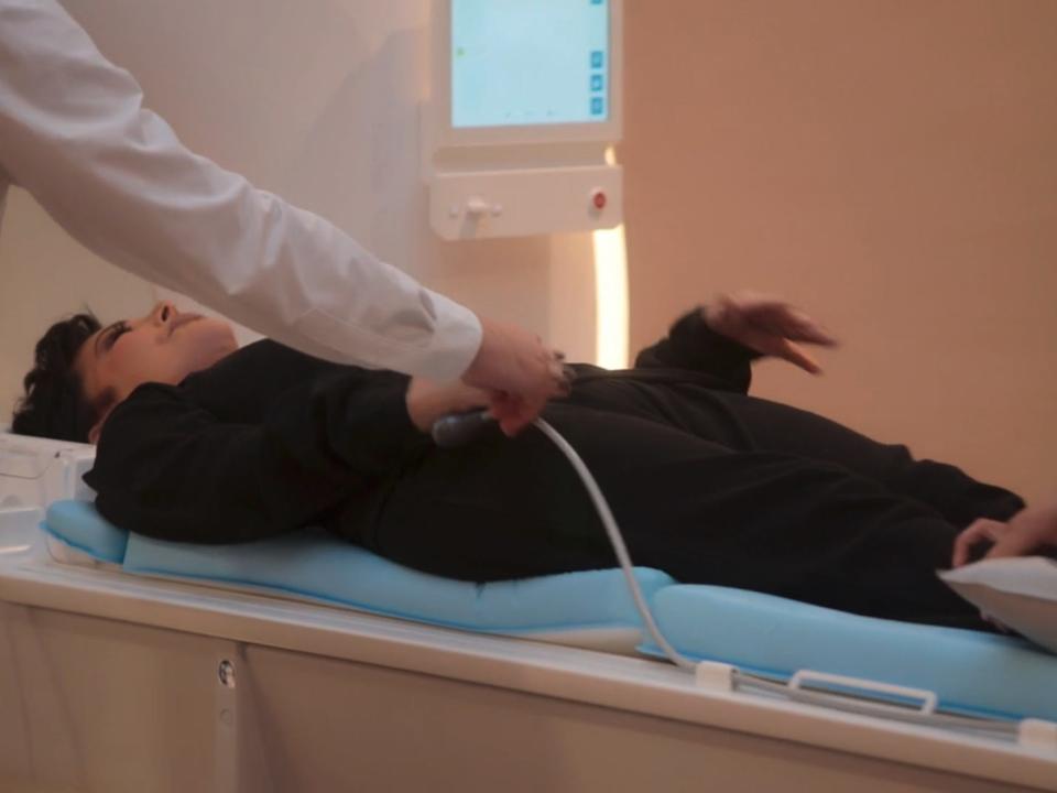 kris jenner laying on a sliding MRI table. there are two techs positioning her on the platform, handing her something to hold in her hand and grabbing a cover to place on top of her body