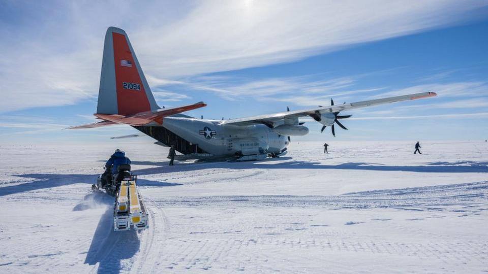 A scientist on a a snow mobile takes a sledge full of drilling equipment towards a US LC-130 Hercules aircraft on Greenland's ice cap