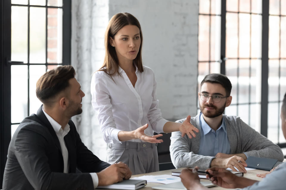Attractive young mentor businesswoman conducts coaching in boardroom at company meeting. Confidence woman training corporate team at briefing. Young employee share thoughts standing at table.