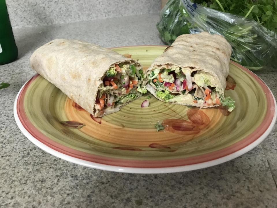 A plate with a halved wrap filled with vegetables and what appears to be avocado, onions, and other fresh ingredients. A bag of greens is in the background