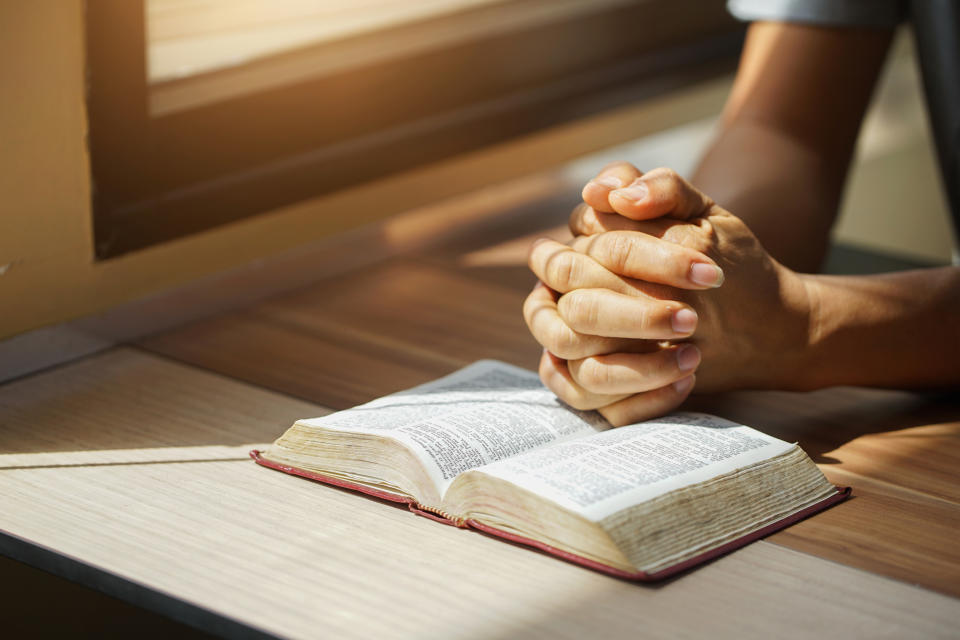 Person's hands clasped together resting on an open book, positioned on a wooden table, with sunlight filtering through a nearby window