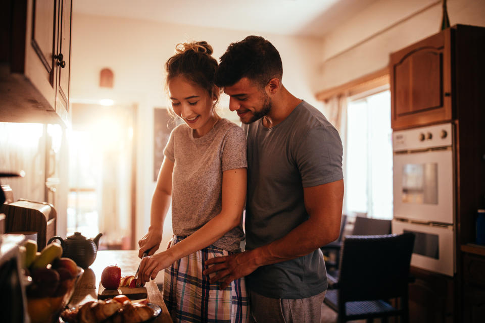 I don't know who these people are. Couple in a cozy kitchen, smiling and preparing food together. The man hugs the woman from behind as she slices bread