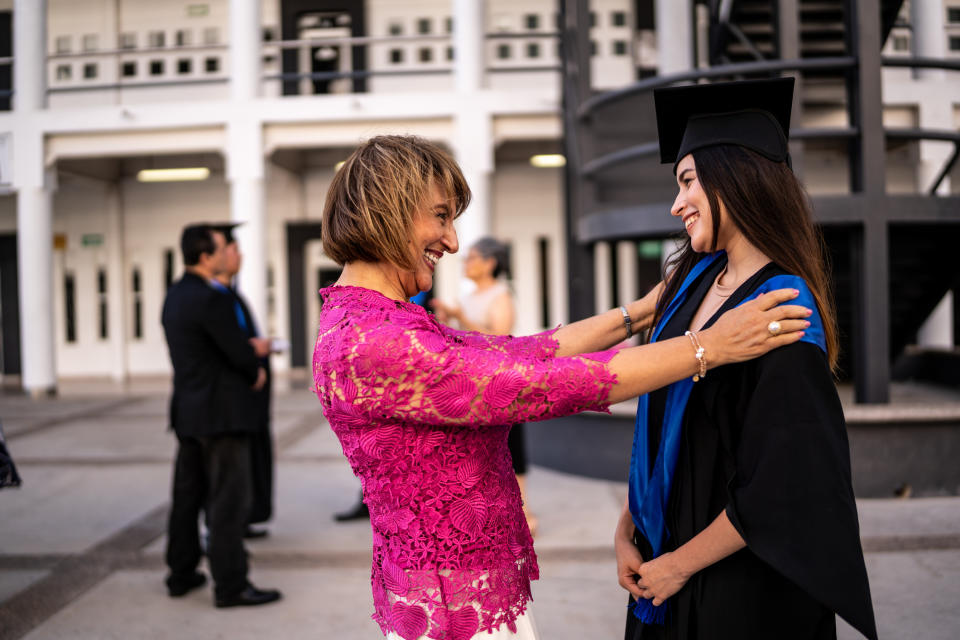Woman in graduation cap and gown smiling at an older woman adjusting her gown, both sharing a happy moment