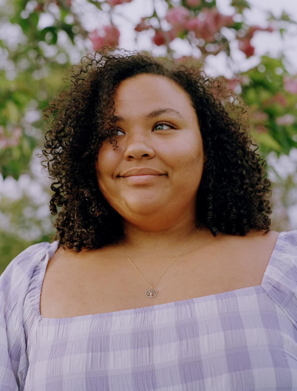 A woman stands in front of a tree wearing a purple checkered dress