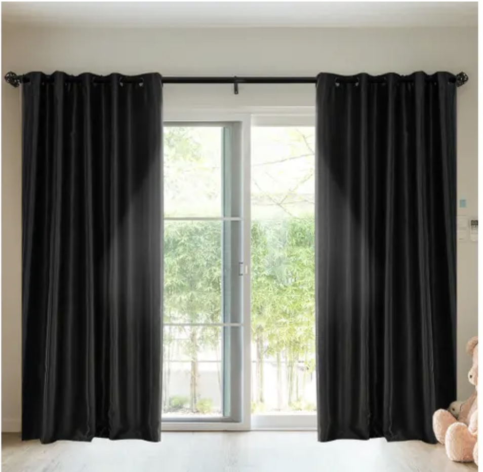 An interior scene looking out through glass onto a garden with black floor-length curtains partially drawn on a white rod. 