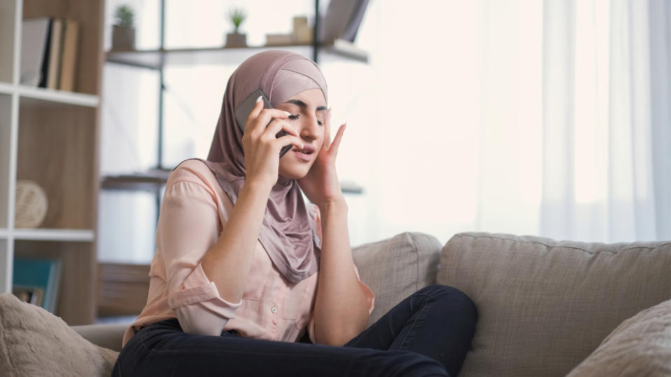 A woman in a headscarf sits on a couch, talking on her cellphone and gesturing with her hand. She appears to be in a living room