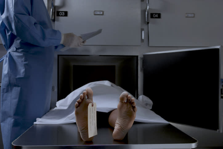 A person lying on a morgue table with a tag on their toe while a medical professional in scrubs and gloves stands next to lockers, holding a clipboard