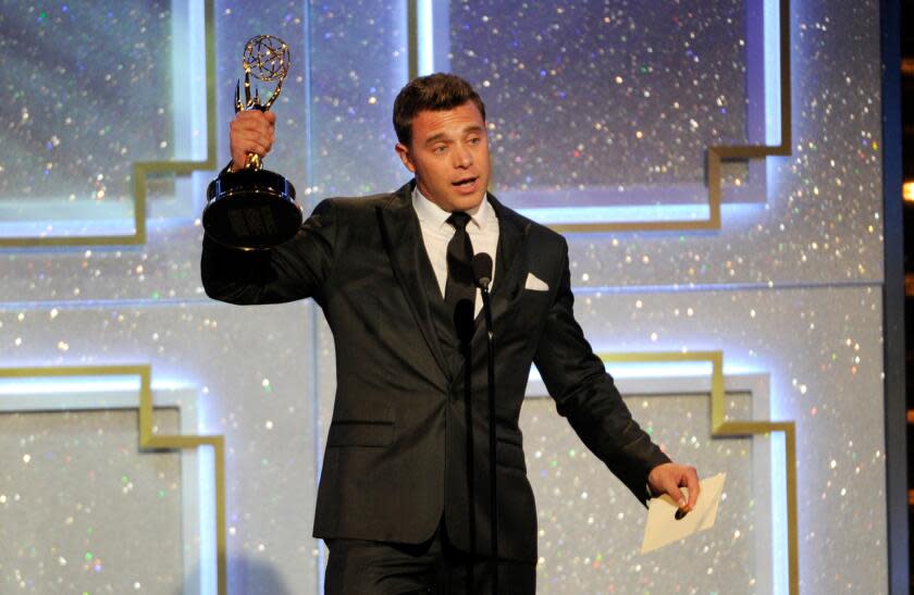 Billy Miller holds up an Emmy Award and an envelope while wearing a black suit and speaking into a microphone on a stage.