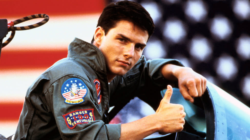  Tom Cruise as Maverick, a pilot in the movie Top Gun, one of the best Prime Video movies, putting up his thumb to the camera as he's leaning on a plane with a blurry United States flag in the background. 