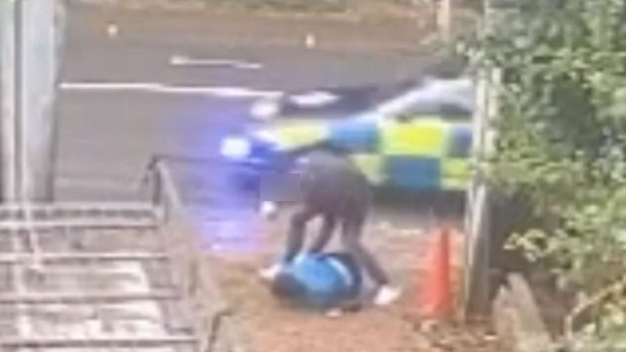 CCTV footage shows a man in dark clothing, his right fist raised, crouching over a man in a blue jacket, face down on the ground. There is a wooden barrier to the right and a police car driving past on the road behind